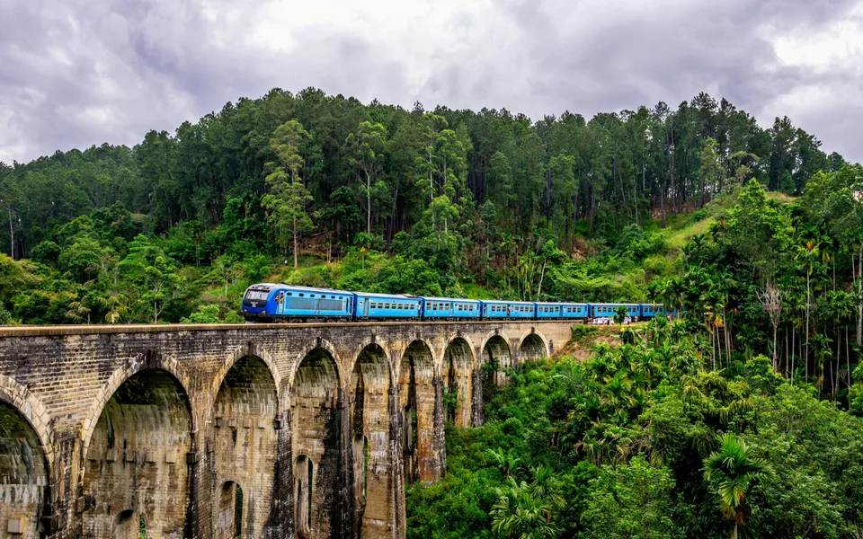 A blue train crossing the Nine Arch Bridge amid green forest in Ella